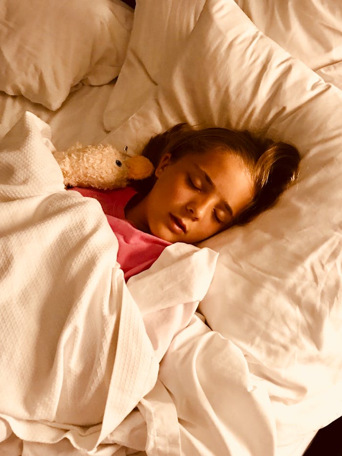 about-us Young girl sleeping peacefully with stuffed animal in a cozy white bed.