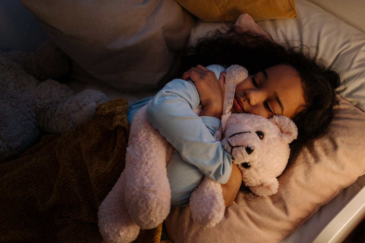 services-03 A young girl peacefully sleeping, hugging a teddy bear in her bed with soft pillows and blankets.